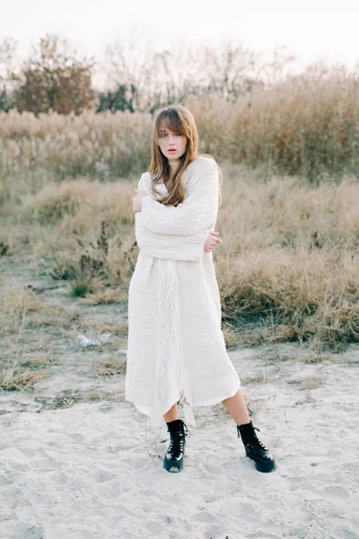 portrait of a young girl in a white dress, a warm knitted cardigan and black shoes among dry reeds and green bushes on the Bank of a river with white sand