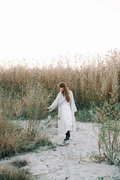 portrait of a young girl in a white dress, a warm knitted cardigan and black shoes among dry reeds and green bushes on the Bank of a river with white sand