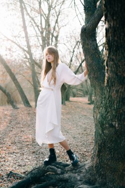 Açık beyaz elbiseli ve siyah ayakkabılı çekici sarışın bir kızın portresi bir ağacın kökünde duruyor.
