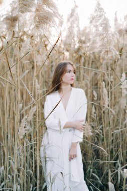 Beyaz elbiseli güzel bir kızın portresi, kalın bir hırka ve kuru kamışların arasında siyah ayakkabılar.