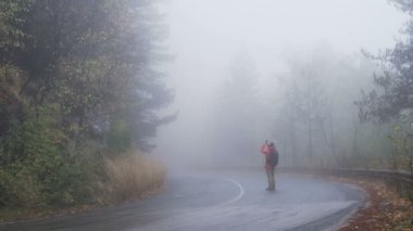 Yalnız turist sisli ormanın telefonuyla fotoğraf çekiyor, boş asfalt yolda duruyor.