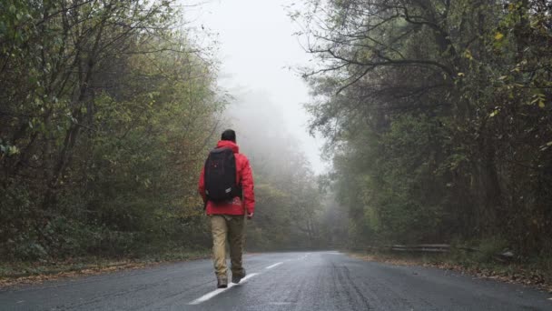 Zorgeloze toerist met rood jasje rustig wandelend op lege mistige weg — Stockvideo
