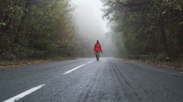 Jovem com casaco vermelho e mochila andando na estrada vazia e abandonada na floresta — Vídeo de Stock