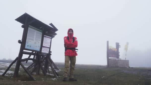 Zamarzający młody człowiek stojący obok drewnianego znaku na szczycie góry w gęstej mgle — Wideo stockowe