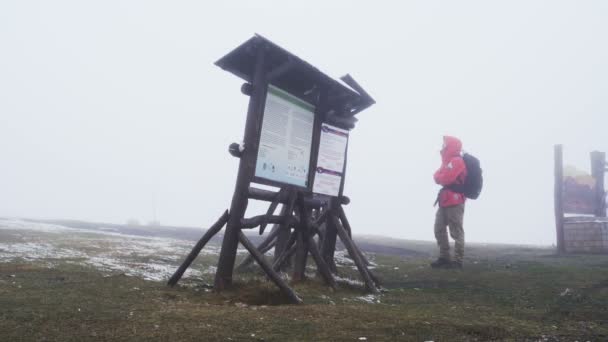 Männlicher Tourist mit Kapuze versteckt sich vor dem Regen und liest Holzschild — Stockvideo