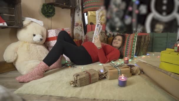 Piękna kobieta w ciąży w czerwonym swetrze świątecznym kłamie i czyta książkę — Wideo stockowe