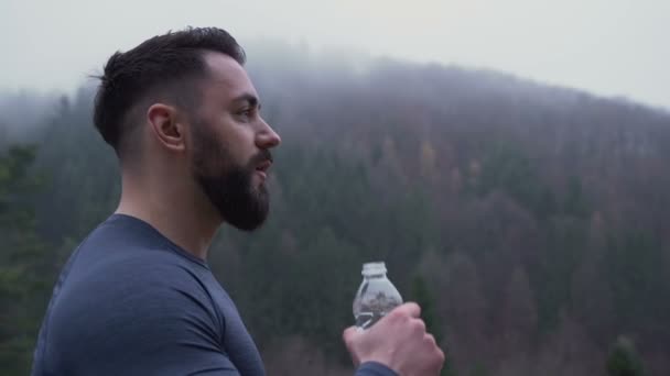 Вид сбоку сильного мужчины с большой прохладной бородой, пьющего воду снаружи на фоне туманного леса — стоковое видео