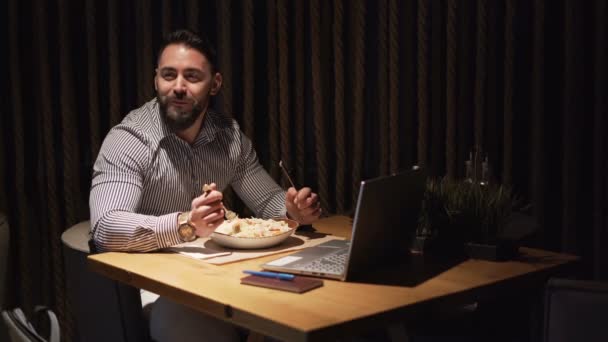 Jonge zakenman eet diner in restaurant met zijn laptop open voor hem, glimlachend — Stockvideo