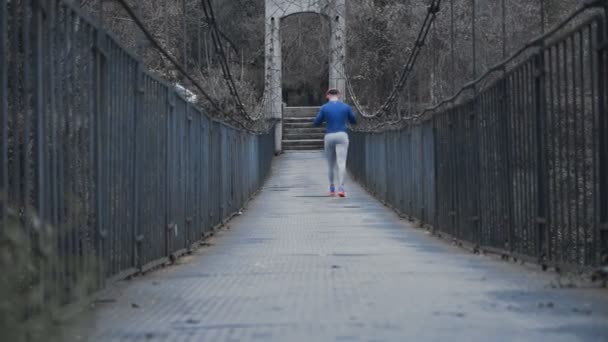 Starker männlicher Läufer mit großen roten Kopfhörern und blauem Hemd, der langsam auf einer Metallbrücke geht — Stockvideo