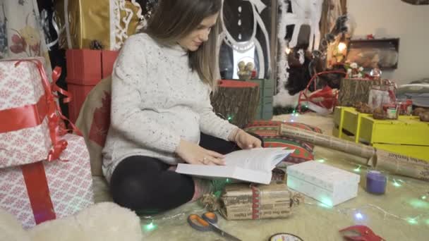 Рождественские огни сияют вокруг беременной женщины. Молодая женщина читает книгу в тёплом свитере — стоковое видео
