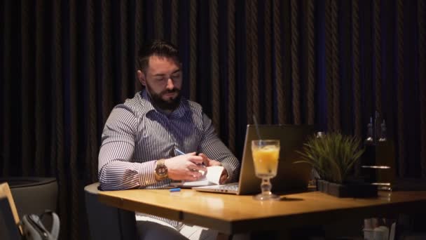 Empresario barbudo tomando notas con pluma mientras trabaja en casa en el ordenador portátil — Vídeos de Stock