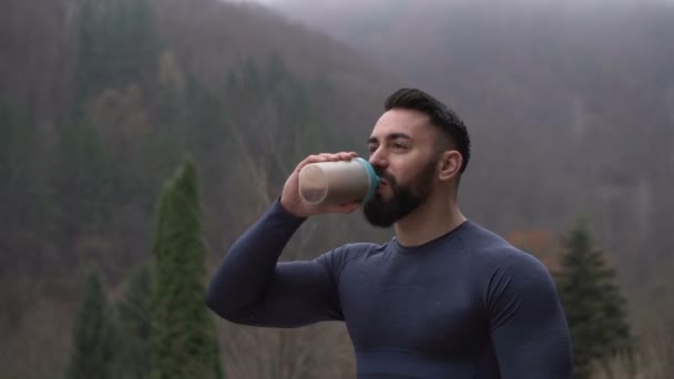 Gezonde sterke man met baard drinken eiwit drinken buiten met mistig bos op de achtergrond — Stockvideo