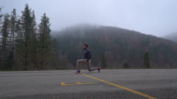 En stilig stark man med atletisk kropp gör stretching övningar för ben på en naturlig bakgrund — Stockvideo