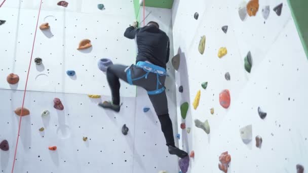 Bergsteiger in einer Boulderhalle. Mann klettert künstliche Mauer hinauf. bunte Bände und Griffe an einer weißen Wand. — Stockvideo