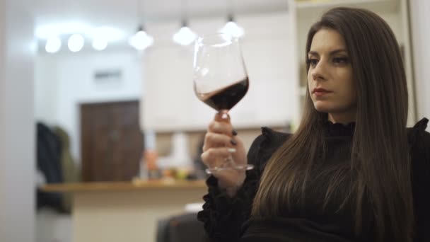 Όμορφη γυναίκα κοιτάζει ένα ποτήρι κρασί, το κουνάει αργά και πίνει μια γουλιά. — Αρχείο Βίντεο