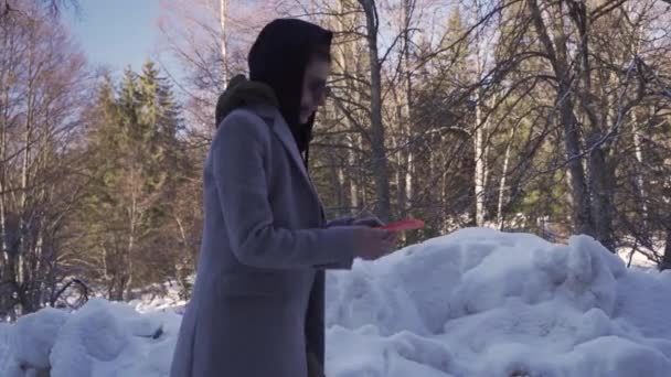 Kapüşonlu ve güneş gözlüklü genç kız karda yürüyor, telefonunu kontrol ediyor. — Stok video
