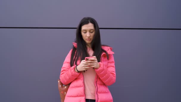 Привлекательная девушка с длинными каштановыми волосами, пишет смс на смартфоне спиной к стене — стоковое видео