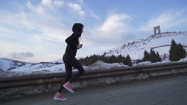 Mujer con sudadera con capucha negra y zapatos rosados de entrenamiento, corriendo en las montañas nevadas — Vídeo de stock