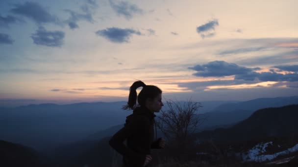 Chica cola de caballo saludando mientras corre en la montaña. Picos de montaña con hermoso cielo al atardecer — Vídeos de Stock