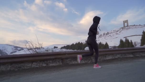Jonge fitnessvrouw joggen in de sneeuw. Arc of Freedom op de achtergrond met kleurrijke wolken bij zonsondergang — Stockvideo