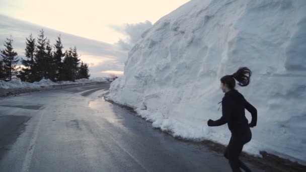 Deportiva mujer con zapatos de color rosa corriendo por el camino de asfalto con grandes pilas de nieve en el lado — Vídeos de Stock