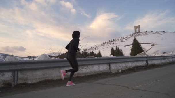 Siguiendo a una joven en ropa deportiva negra trotando en las montañas, alcanzando el Arco de la Libertad en la cima — Vídeo de stock