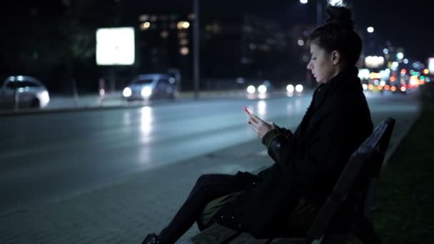 Chica esperando a su amigo en el banco, enviando mensajes de texto a un amigo desde el teléfono inteligente — Vídeo de stock