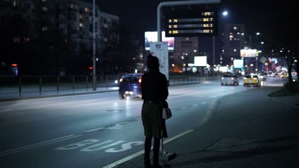Zamarzająca dziewczyna patrzy, czeka na przystanku autobusowym w nocy — Wideo stockowe