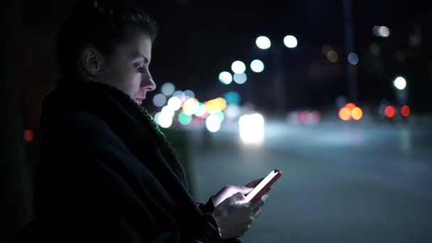 Vista lateral da jovem usando smartphone com cidade noturna em segundo plano — Vídeo de Stock