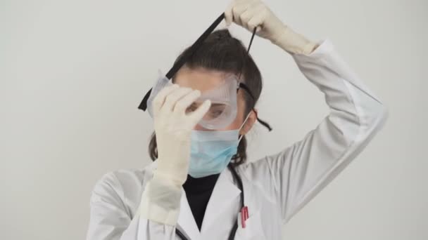 Молодая женщина-врач надевает защитные пластиковые очки и маску против коронавируса — стоковое видео