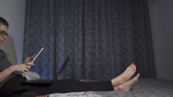 Donna con occhiali sdraiata a letto che lavora con tablet e laptop — Video Stock