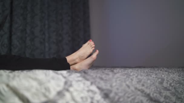 Молодая женщина с босыми ногами и красным лаком для ногтей лежит в постели улыбаясь, используя свой смартфон — стоковое видео