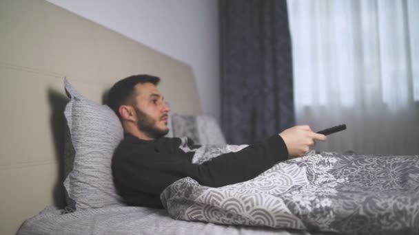 Sick man lying bed watching TV switching channels with remote — Stock Video