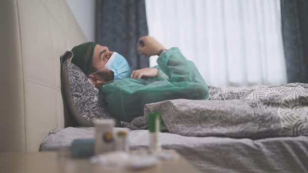 ベッドに横たわっながら、保護服、帽子とフェイスマスクを持つ病気の男は、温度計を取ります — ストック動画