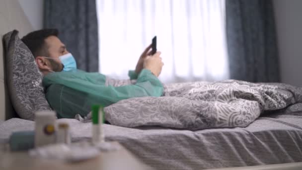 Quarantined man infected with virus lying in bed in protective clothes and reading news on his smartphone — Stock Video