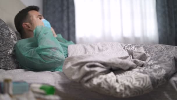 Köhögés férfi védőruha üzembe arc maszk feküdt az ágyban mellett éjjeliszekrény több gyógyszer és tabletta — Stock videók