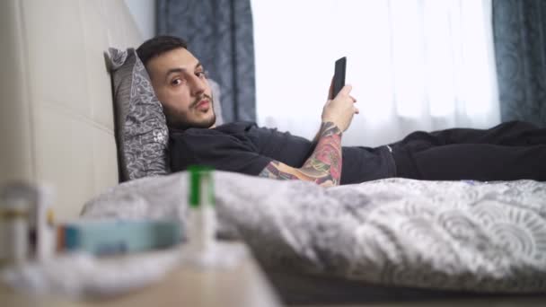 疲れて病気の若い男が横にカメラを見てベッドに横たわって — ストック動画