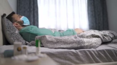 Evde tek başına öksüren adam mavi yüz maskesi ve sağlık kıyafeti giyiyor.