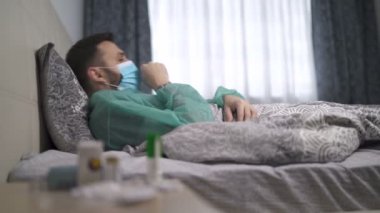 Ateşli, öksüren hasta bir adam evde yatakta yatıyor. Yüz maskesi ve koruyucu sağlık kıyafeti giyen karantina altındaki bir adam.