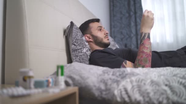 Хворий молодий чоловік з лихоманкою лежить в ліжку і перевіряє температуру за допомогою термометра пахви — стокове відео