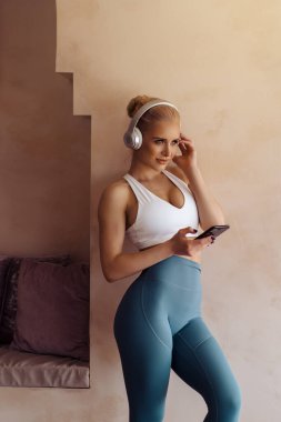 Kulaklıklı, akıllı telefonu elinde tutan güzel ve sağlıklı genç bir kadın. Evde egzersiz yaparken müzik dinleyen çekici, sportif bir kadın..