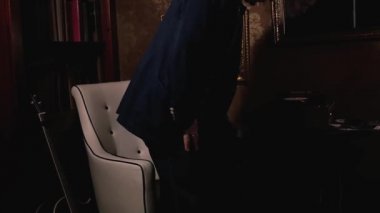 Yaşlı bir adam, kitaplar, tablolar, pikaplar ve diğer sanat eserleriyle dolu büyük bir kütüphanede beyaz deri bir sandalyede oturuyor. Harika bir sanat ve sosyal konsept..