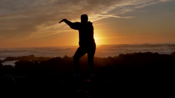 Erkek savaşçı keşiş gün batımında ya da gün doğumunda kayalık kayalıklarda Tai Chi karate kung fu pratik yapıyor. Kendini savunma sanatı. Pasifik sahilindeki dramatik destansı dalgaların arka planında siluet — Stok video