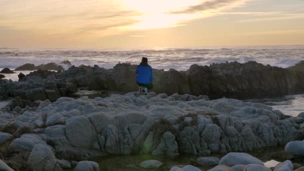 Slow motion cinematic van de mens zitten in blauwe jas en wandelen op rotsachtige kust bij zonsondergang california vibes pacific oceaan — Stockvideo