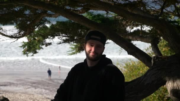 Um close-up retrato em câmera lenta do jovem millennial sentado na árvore sorrindo e olhando para a câmera brincando com a sombra. Dia ondas de luz mar de praia no fundo . — Vídeo de Stock