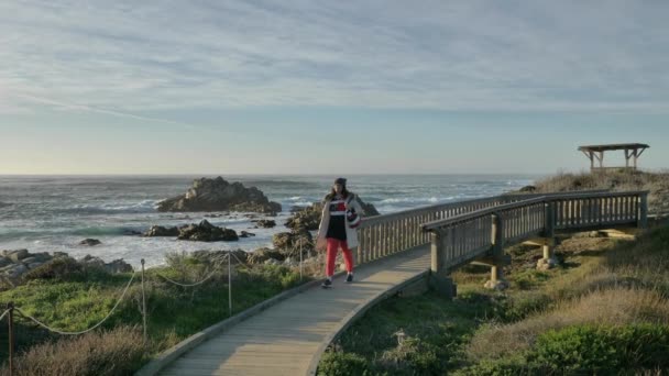 Aktivní starší běloška kráčející po mostě v pacifickém pobřeží s bílým pláštěm a teplým kloboukem, užívající si dobrodružství pomalé pohyby — Stock video