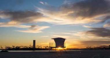 Bulutlar, cankurtaran kulesi ve Long Beach 'te arka planda vinçlerle güzel bir günbatımı okyanusu. Los Angeles, Kaliforniya. 4k Zaman Çizelgesi.
