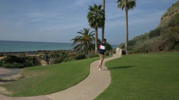 Um cara bonito, jovem, um homem com uma camisa azul marinho e shorts está andando ao longo da costa da Califórnia San clemente Linda Lane Park — Vídeo de Stock