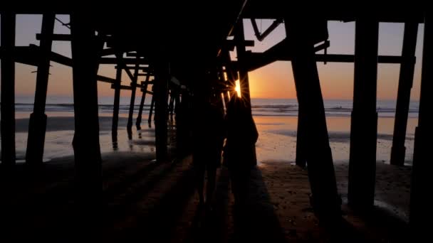 Par gå nära hålla händerna tillsammans på natursköna orange rosa solnedgång med episka strålar av ljus och sol fackla trä piren i San Clementa Kalifornien — Stockvideo