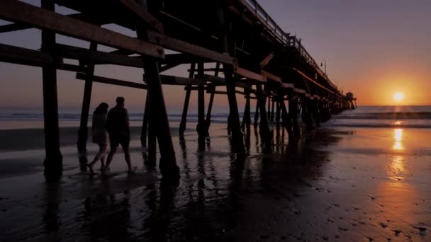 Ζευγάρι περπατώντας μαζί κρατώντας hads Γραφικό πορτοκαλί ροζ Ηλιοβασίλεμα με επικές ακτίνες του φωτός και του ήλιου φωτοβολίδα ξύλινη προβλήτα στο San Clementa Καλιφόρνια — Αρχείο Βίντεο